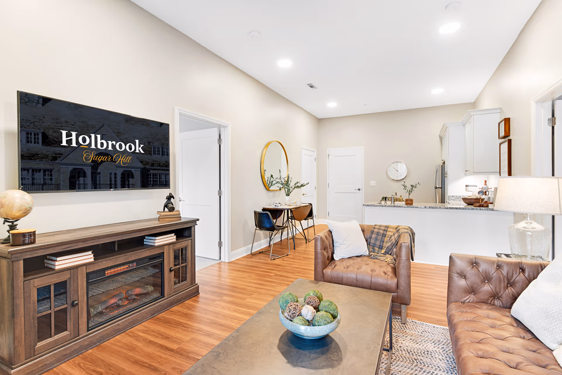 A bright and modern senior living facility interior featuring a cozy living room with two brown leather tufted chairs and a matching sofa, a coffee table with decorative balls, a wooden media console with an electric fireplace, and a wall-mounted TV displaying 'Holbrook Sugar Hill'. The room has light-colored walls, wood flooring, recessed ceiling lights, and an open view into a kitchen area with white cabinetry and granite countertops. A small dining area with two chairs and a round mirror is visible along the wall.