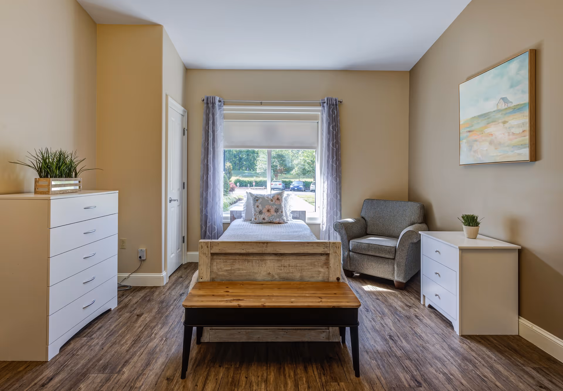 A bright and cozy bedroom with a single bed centered against a window with sheer curtains. The room features wood flooring, a wooden bench at the foot of the bed, a gray armchair to the right, a white dresser with a small plant on top on the right side, and a taller white chest of drawers with a plant on the left side. A framed landscape painting hangs on the right wall.
