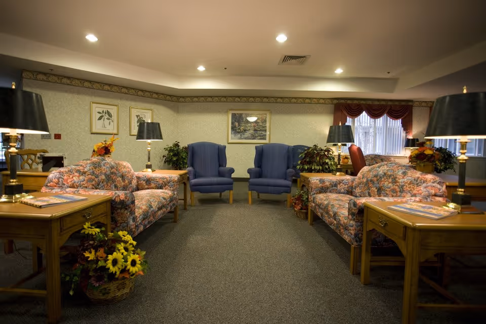 A cozy living room area in a senior living facility with two floral patterned sofas facing each other, two blue armchairs against the far wall, wooden side tables with black lamps, and decorative plants and flowers. The walls are adorned with framed artwork and the room is softly lit with ceiling lights.