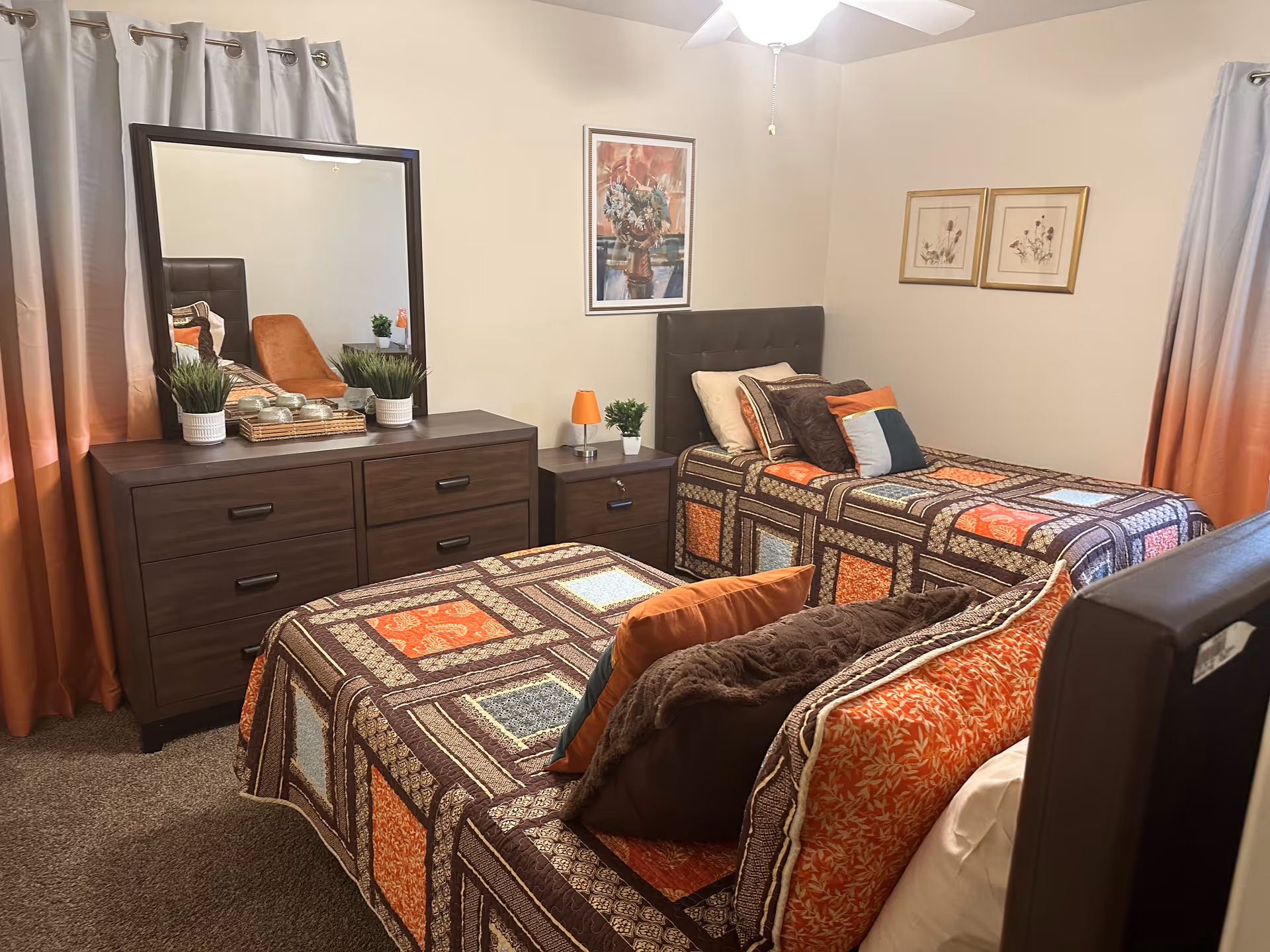 A bedroom with two twin beds featuring colorful patchwork quilts in shades of orange, brown, and blue. Each bed has multiple pillows. Between the beds is a small nightstand with a small orange lamp and a potted plant. Against the wall is a dark wooden dresser with a large mirror on top, decorated with two potted plants and a tray with candles. The room has beige walls with framed artwork and orange and gray ombre curtains on the windows.