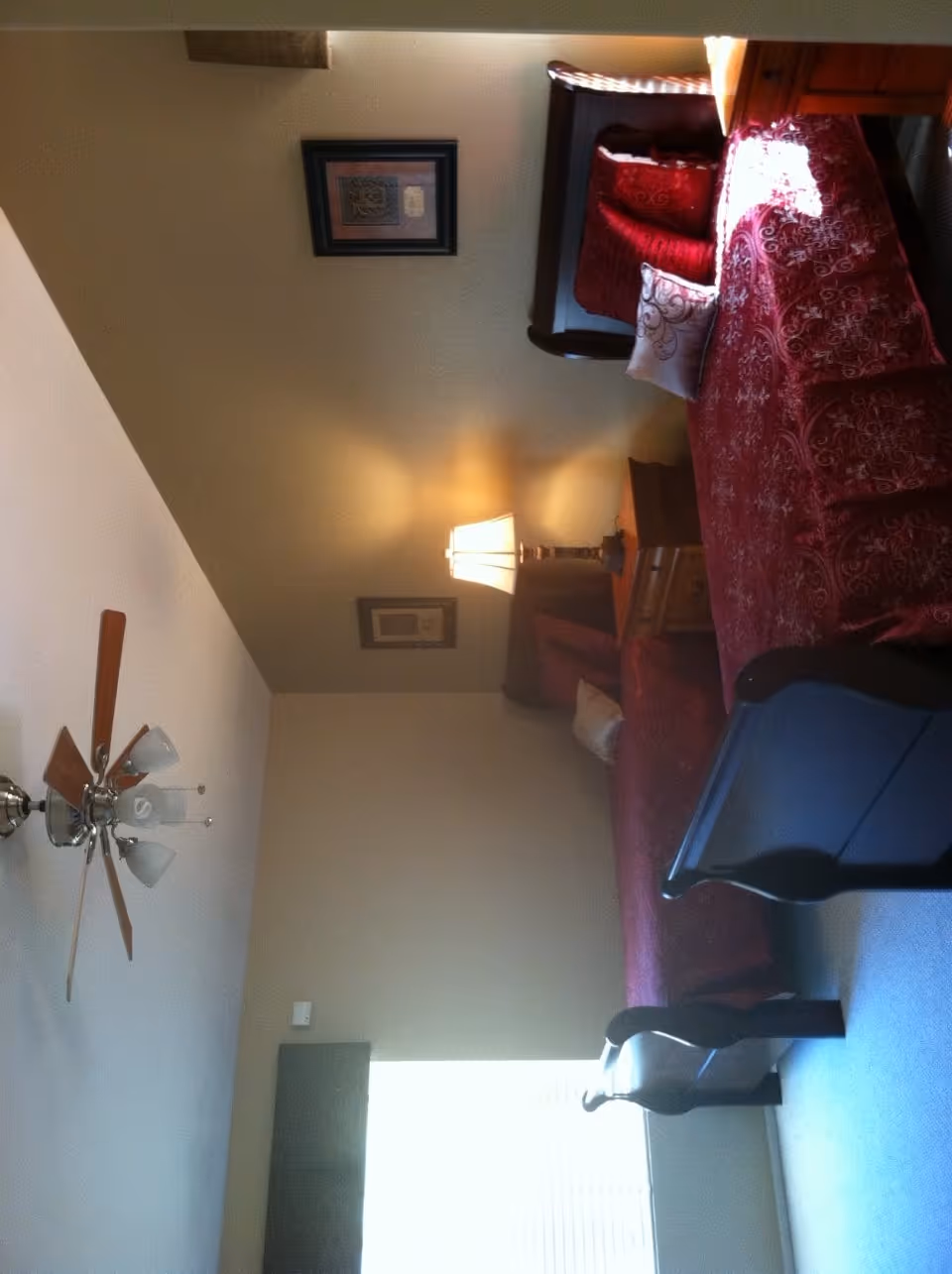 A bedroom with a bed covered in a red patterned bedspread and matching pillows. There is a wooden nightstand with a lamp emitting warm light. The walls are beige with two framed pictures hanging. A ceiling fan with lights is visible on the ceiling, and a window with blinds is partially visible.