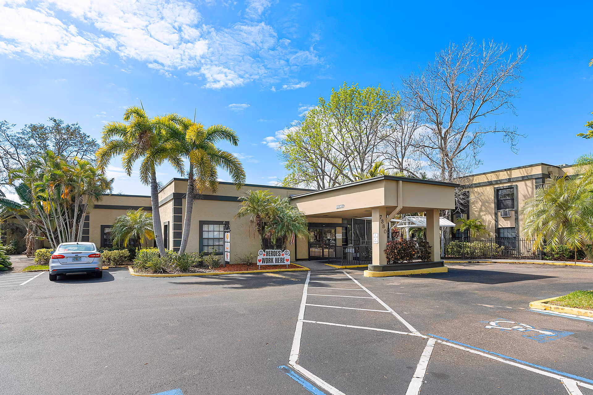 Exterior view of Tierra Pines Center showing a single-story building with a covered entrance, surrounded by palm trees and other greenery under a blue sky with scattered clouds. A white car is parked in the lot, and a sign near the entrance reads 'Heroes Work Here'.