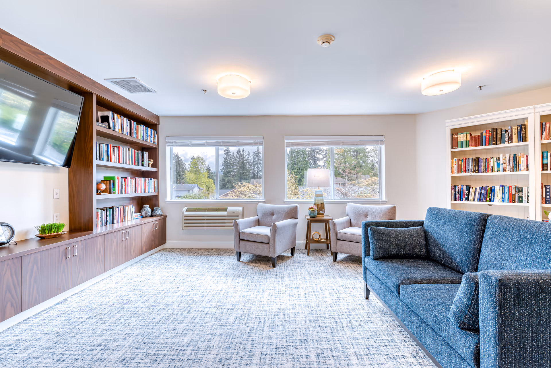 A bright and spacious living room with large windows showing trees outside. The room features a blue sofa, two gray armchairs with a small table and lamp between them, built-in wooden bookshelves filled with books, a mounted flat-screen TV, and a patterned carpet.