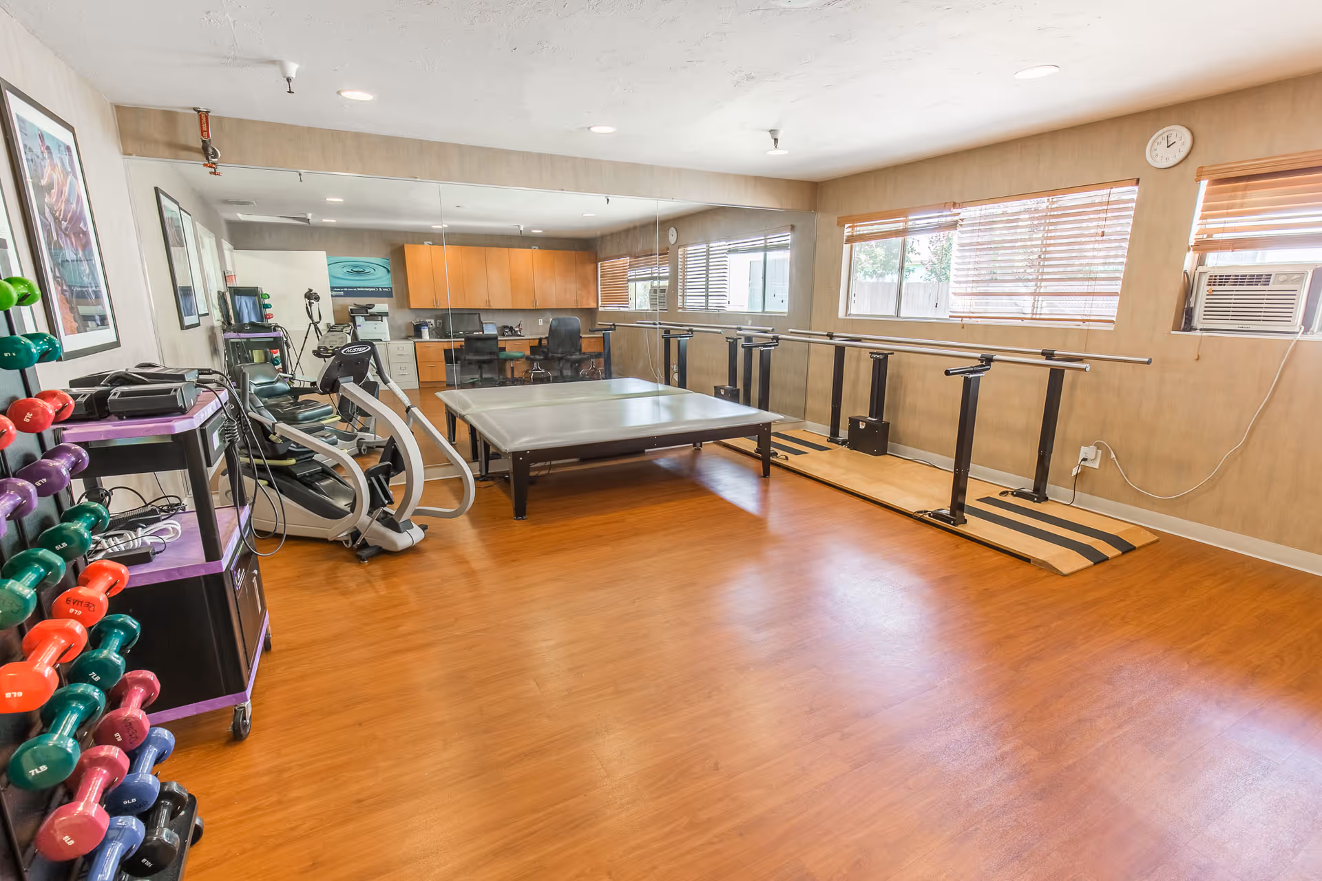 A bright exercise/therapy room with wood floors, mirrored wall, parallel bars, a therapy table, cardio equipment and a rack of colorful dumbbells.