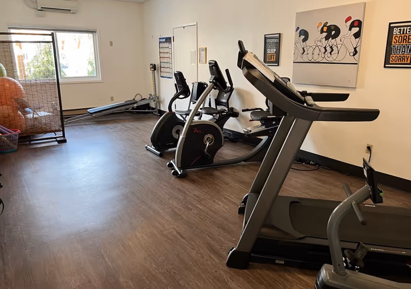A fitness room with exercise equipment including a treadmill, stationary bikes, and a rowing machine. The room has wood flooring, white walls with motivational posters, a window letting in natural light, and a basket with large exercise balls in the corner.