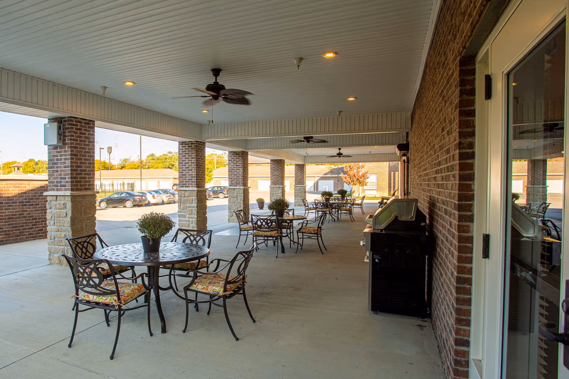Covered outdoor patio area with multiple round metal tables and chairs with floral cushions, potted plants on the tables, ceiling fans, and a grill against a brick wall. In the background, there is a parking lot with cars and some buildings.