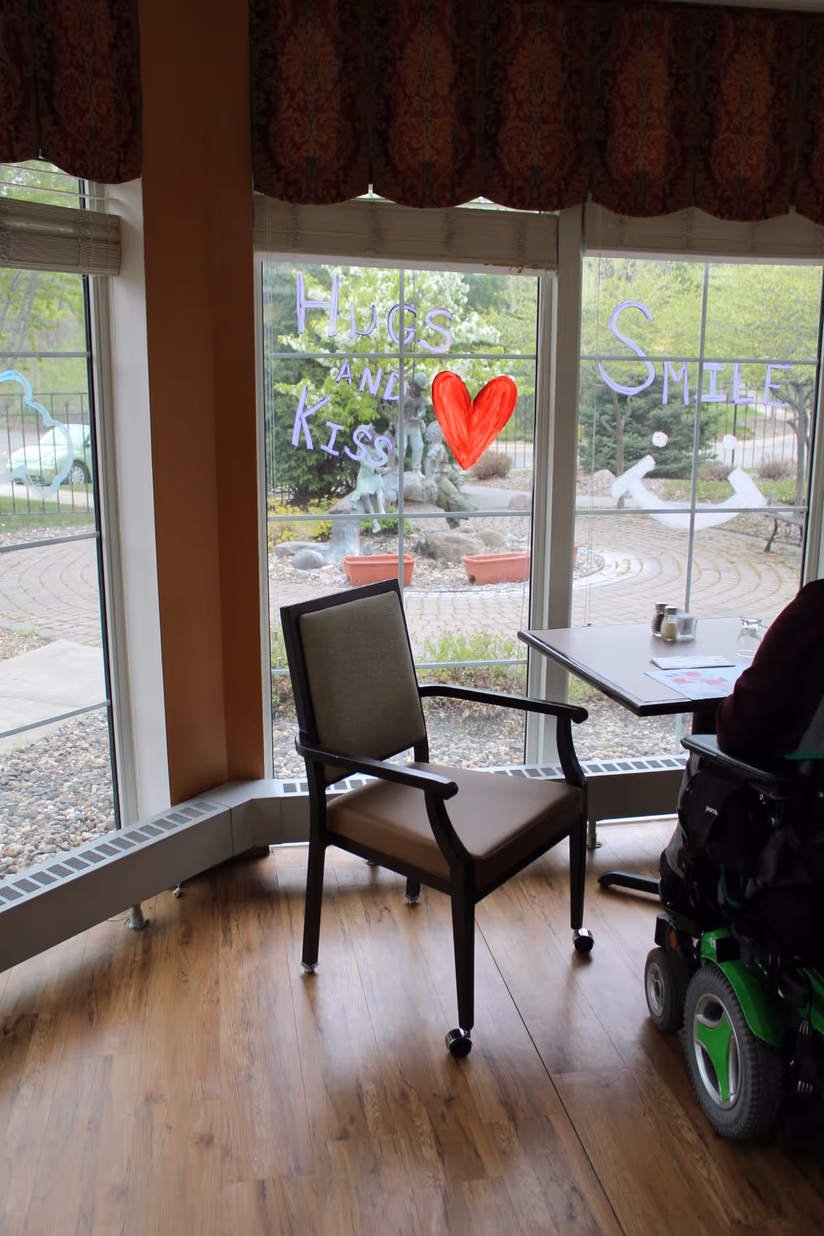 A corner seating area with a single chair and a table next to large windows. The windows have colorful writing on them that says 'Hugs and Kiss' with a red heart and 'Smile' with a smiley face. Outside the windows, there is a paved circular garden area with plants and a sculpture.