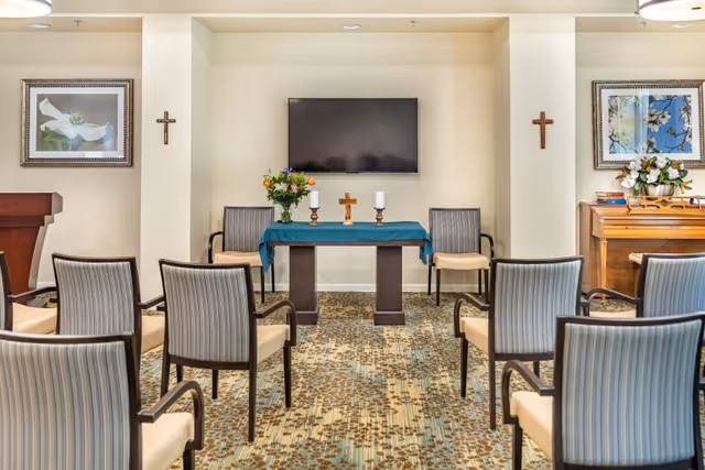 A small chapel or prayer room with rows of chairs facing a table covered with a blue cloth, holding a wooden cross, two candles, and a vase of flowers. The room has a flat-screen TV mounted on the wall, two framed floral paintings, two wooden crosses on the walls, and a piano with flowers and books on top.