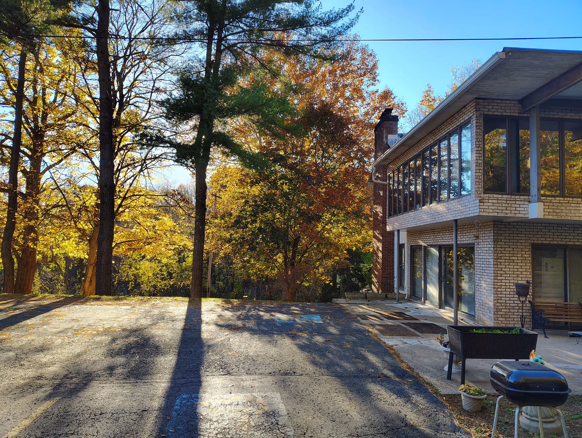 Exterior view of a two-story brick building with large windows, surrounded by trees with autumn foliage. There is a paved area in front with shadows from the trees, a black grill, a planter, and a bench near the building.