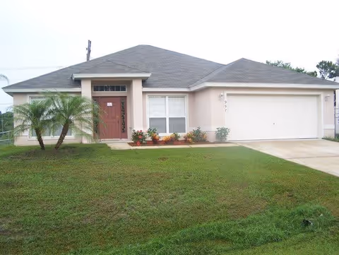 Single-story residential building with a gray roof, beige exterior walls, a red front door, two windows, a two-car garage, and a small landscaped area with plants and a palm tree in front.