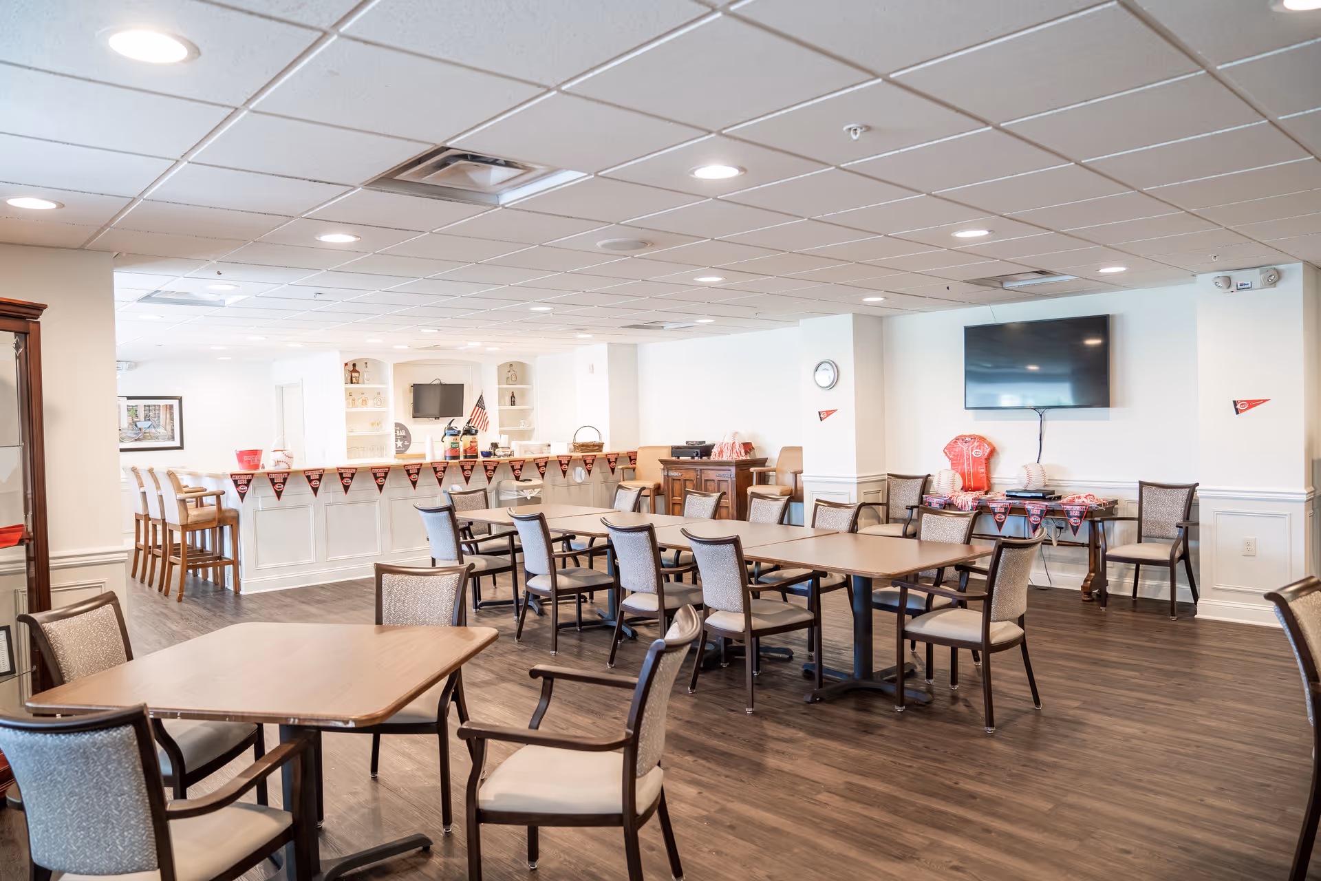 A spacious dining room with multiple tables and chairs arranged neatly on a wooden floor. There is a counter area in the back with bar stools and shelves holding various items. The room is decorated with sports-themed banners and memorabilia, including a red jersey and baseballs displayed on a table beneath a wall-mounted TV. The ceiling has recessed lighting, and the walls are painted white.