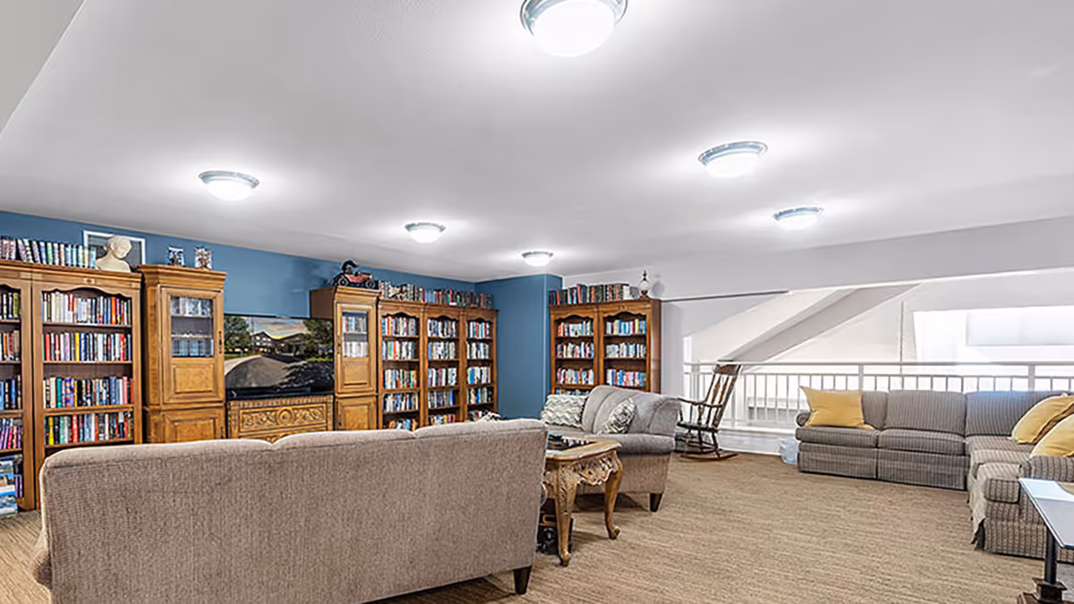 A spacious living room area with multiple bookshelves filled with books along a blue wall. The room features several comfortable sofas and armchairs arranged around a wooden coffee table. The ceiling has multiple round light fixtures, and there is a rocking chair near a white railing with large windows letting in natural light.