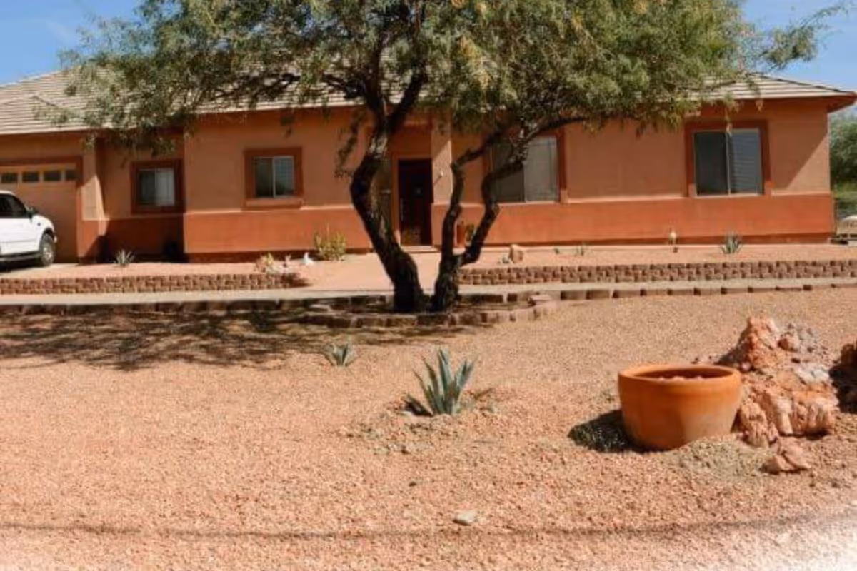 Single-story building with a tan and reddish exterior, a tree in front, gravel landscaping with small plants, a large clay pot, and a white vehicle parked on the left side.