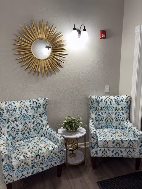 Two patterned armchairs with blue and beige designs are placed on either side of a small round table with a potted plant. Above the chairs, there is a decorative sunburst mirror and a wall-mounted light fixture with two bulbs. The wall is painted light gray and there is a fire alarm and light switch visible.