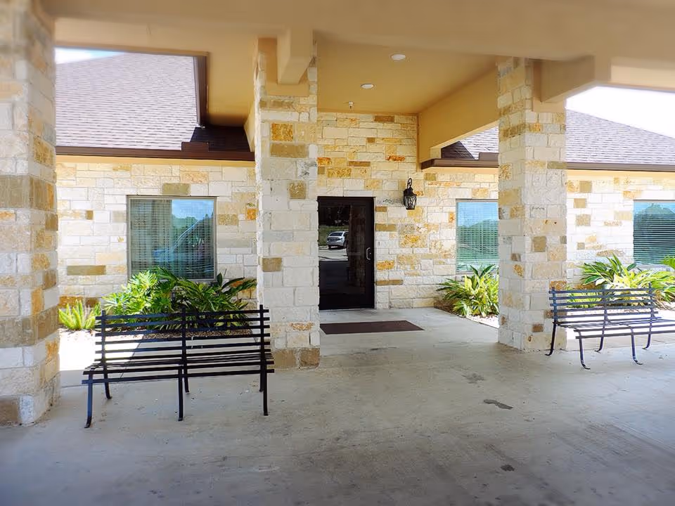 Covered entrance area of a building with stone walls, two black metal benches, potted plants, and a glass door in the center. The area is shaded by a roof supported by stone pillars.