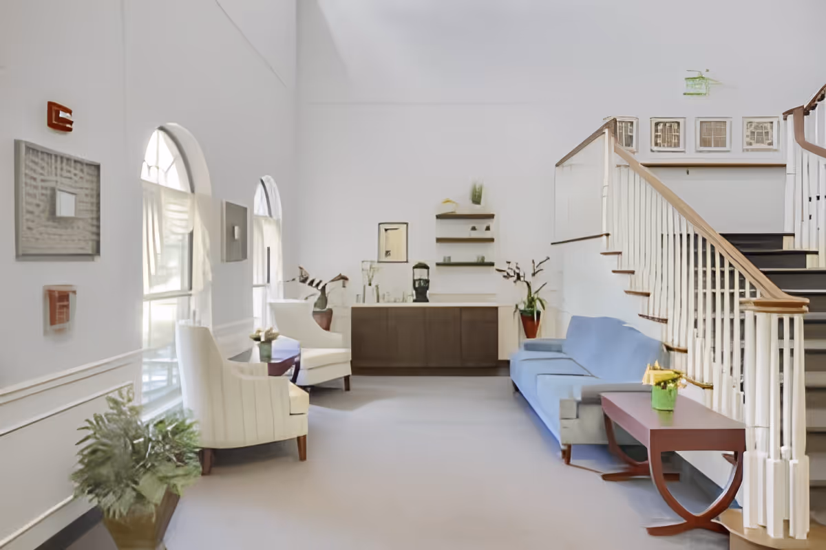 Bright seating area in a senior living facility lobby with chairs, a blue sofa, side tables, plants, and a staircase.
