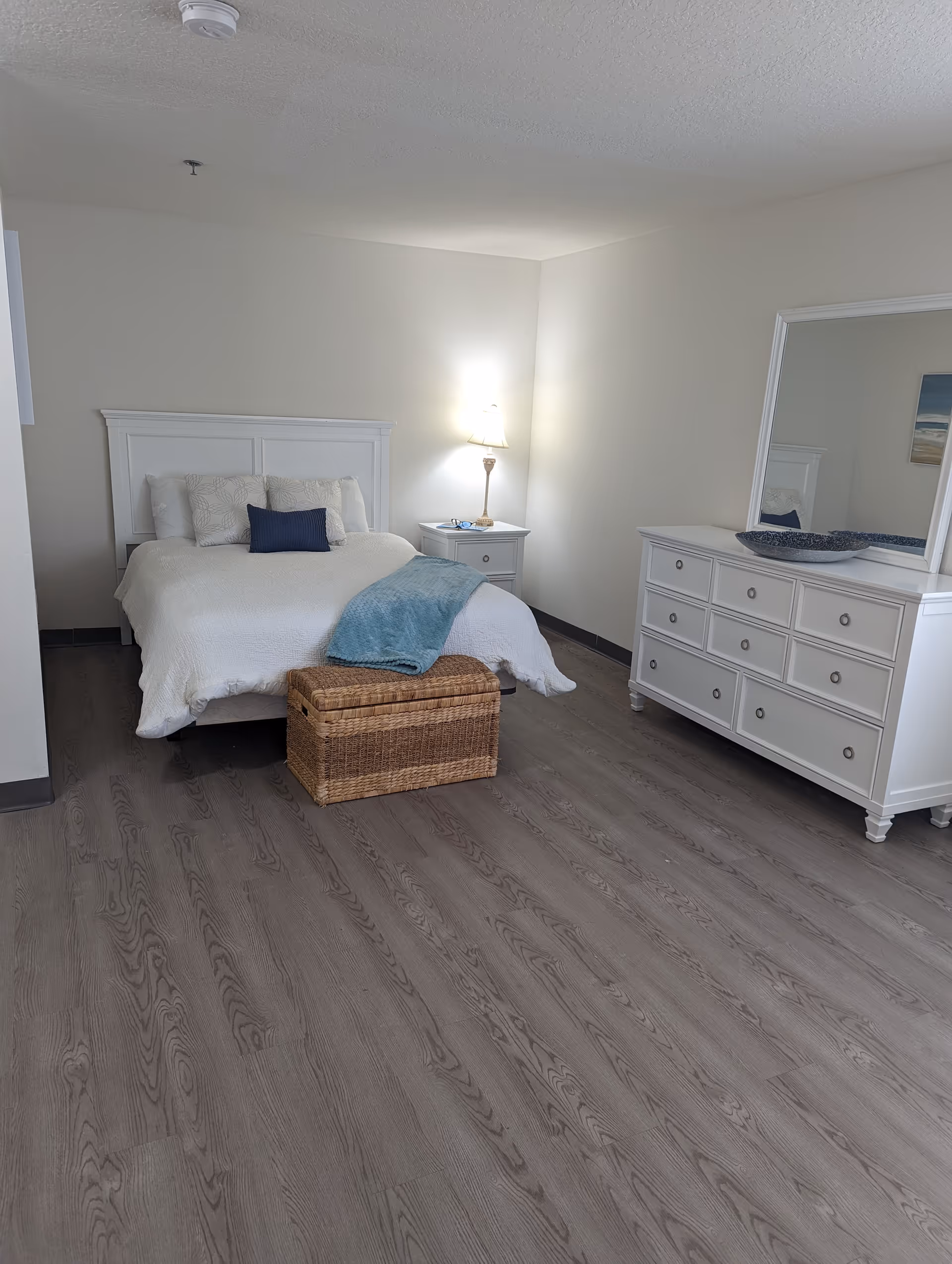 A clean and simple bedroom with a white bed featuring white and blue pillows, a blue throw blanket, a wicker storage chest at the foot of the bed, a white nightstand with a lamp, and a white dresser with a large mirror and decorative bowl on top. The room has light-colored walls and wood flooring.