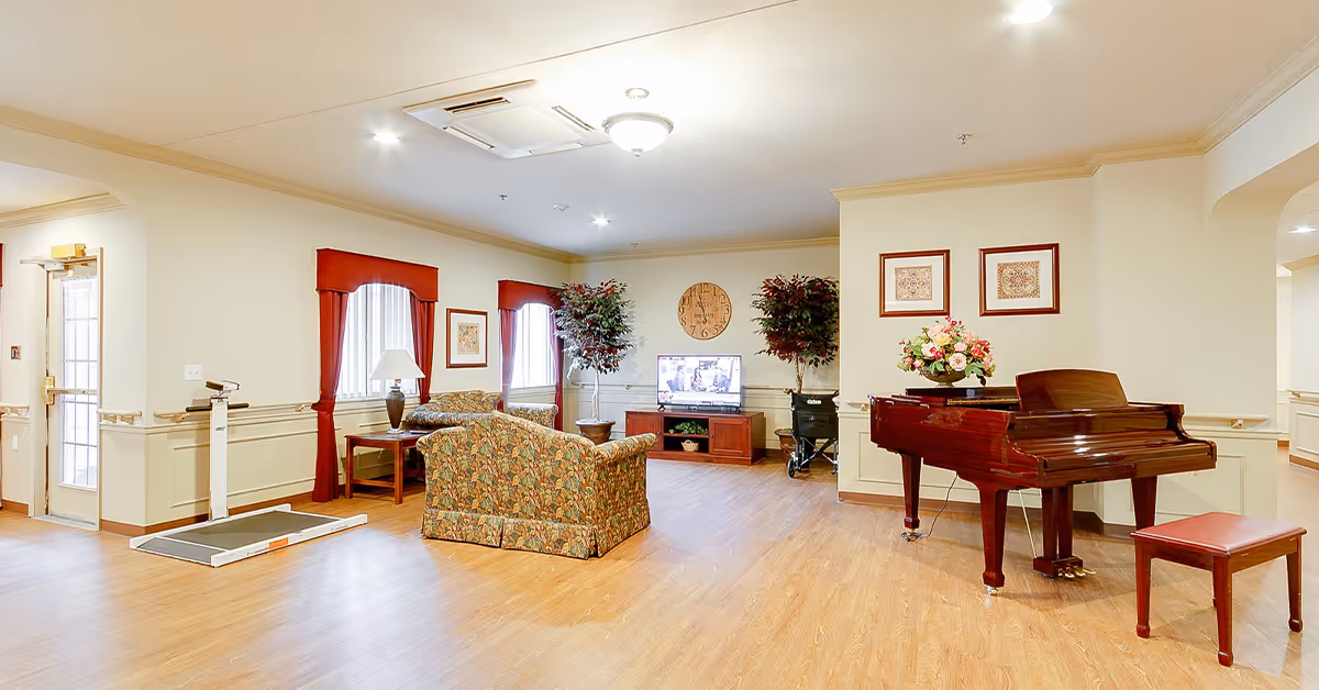 A bright and spacious senior living common area with wooden flooring, a floral-patterned sofa and armchair, a wooden coffee table with a lamp, two large potted plants, a flat-screen TV on a wooden stand, a large wall clock, a grand piano with a matching bench, and framed artwork on the walls. There is a door with a ramp on the left side and arched doorways leading to other rooms.