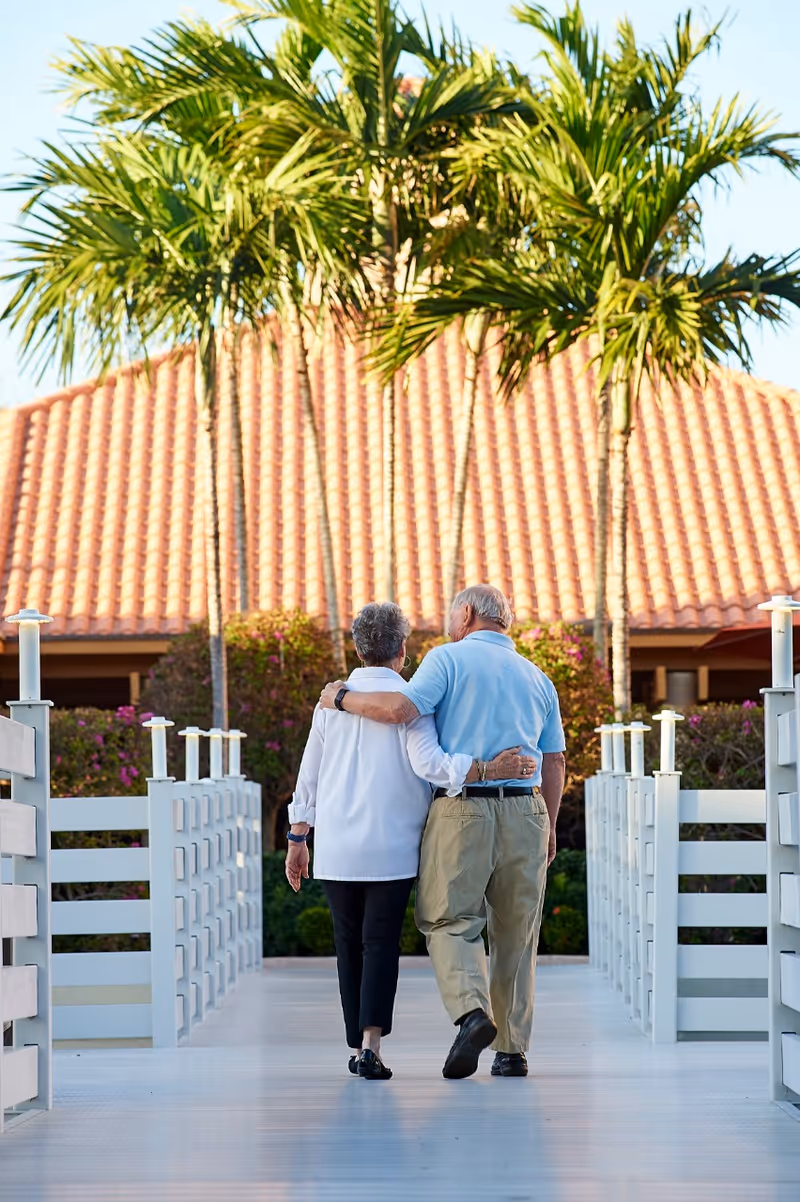 An elderly couple walking arm in arm on a white wooden bridge with palm trees and a building with a red tiled roof in the background.