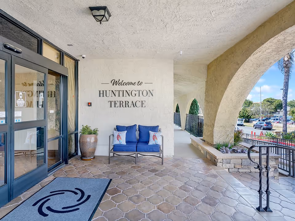 Entrance area of Huntington Terrace facility with a welcome sign on the wall, a blue cushioned bench with decorative pillows, a large potted plant, tiled floor, and arched openings showing a parking lot and greenery outside.