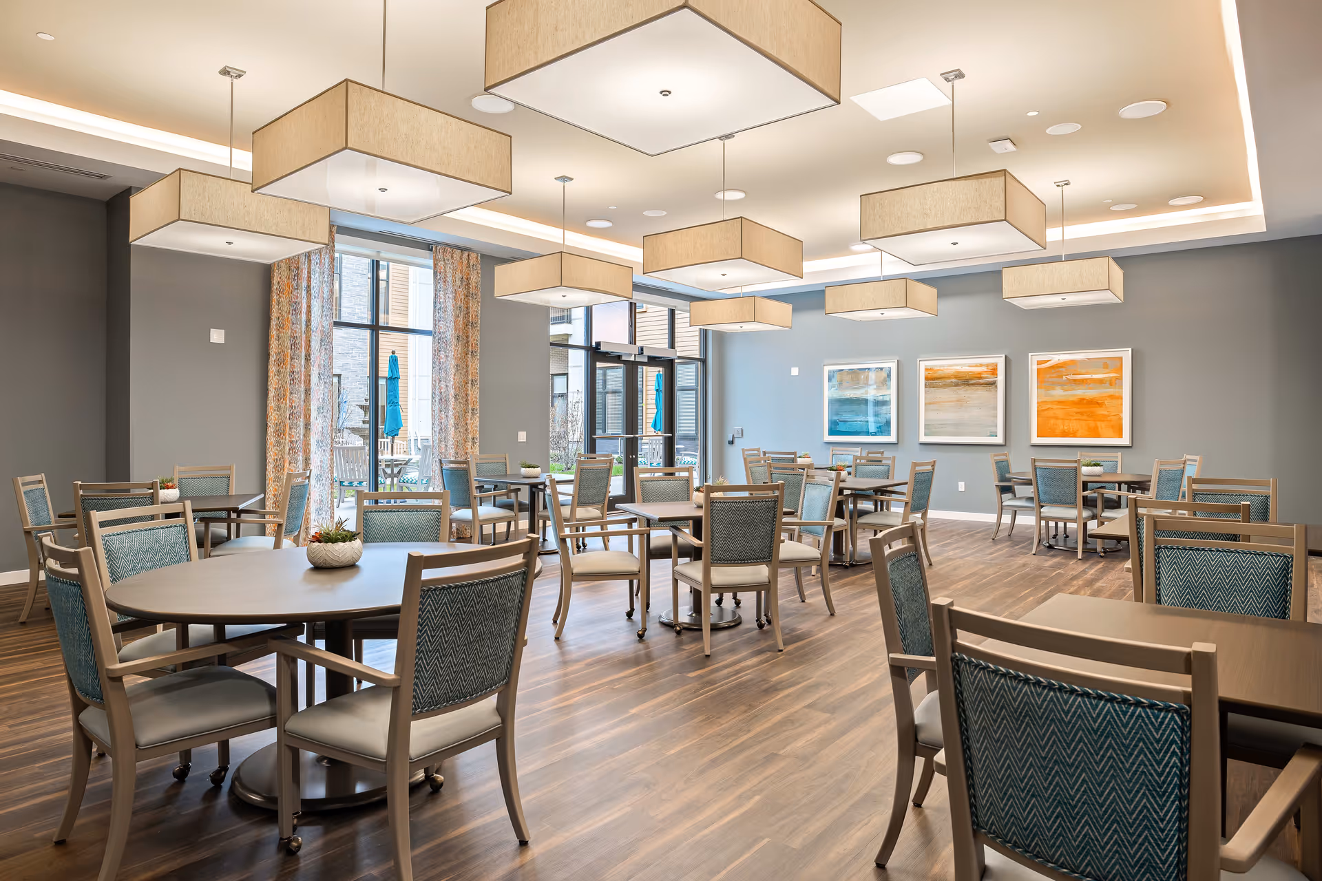 A spacious dining room with multiple tables and chairs arranged neatly on a wooden floor. The room features large windows with patterned curtains allowing natural light to enter. Modern rectangular ceiling lights hang above the tables. Three abstract paintings in blue, gray, and orange tones are displayed on a gray wall.