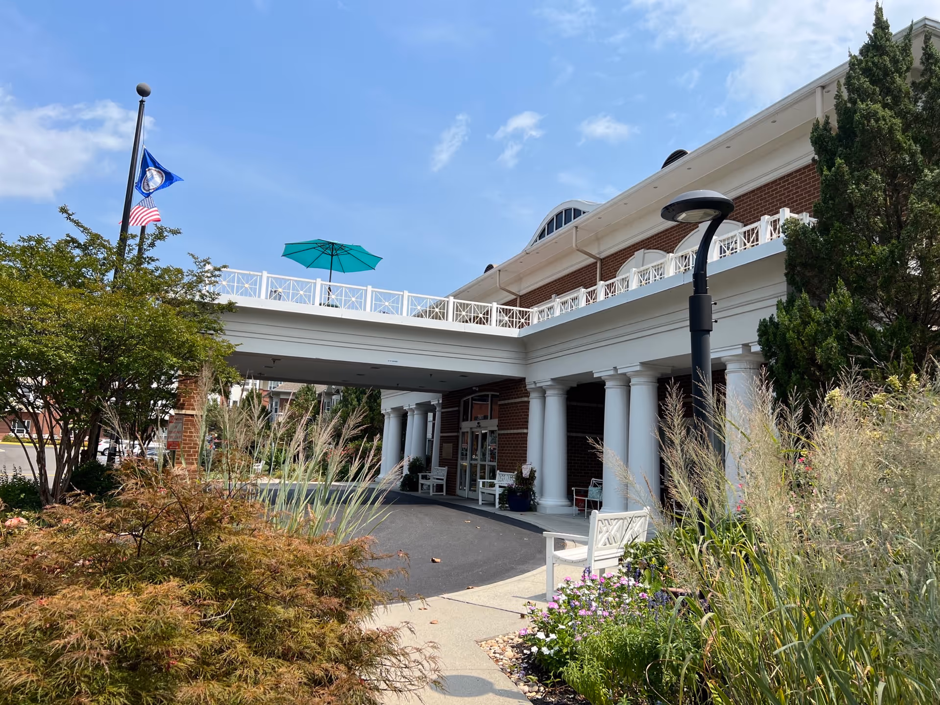 Exterior view of Shalom Gardens Senior Assisted Living Facility in Virginia showing the entrance with white columns, a covered driveway, landscaped garden with flowers and shrubs, a street lamp, and flags flying on a flagpole against a blue sky.