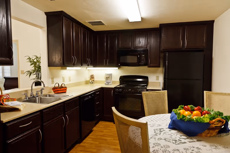 A kitchen with dark wood cabinets, a double sink, black appliances including a stove, microwave, dishwasher, and refrigerator. There is a round dining table with a white lace tablecloth and a basket of assorted fresh fruits and vegetables in the foreground. The kitchen has under-cabinet lighting and a wooden floor.