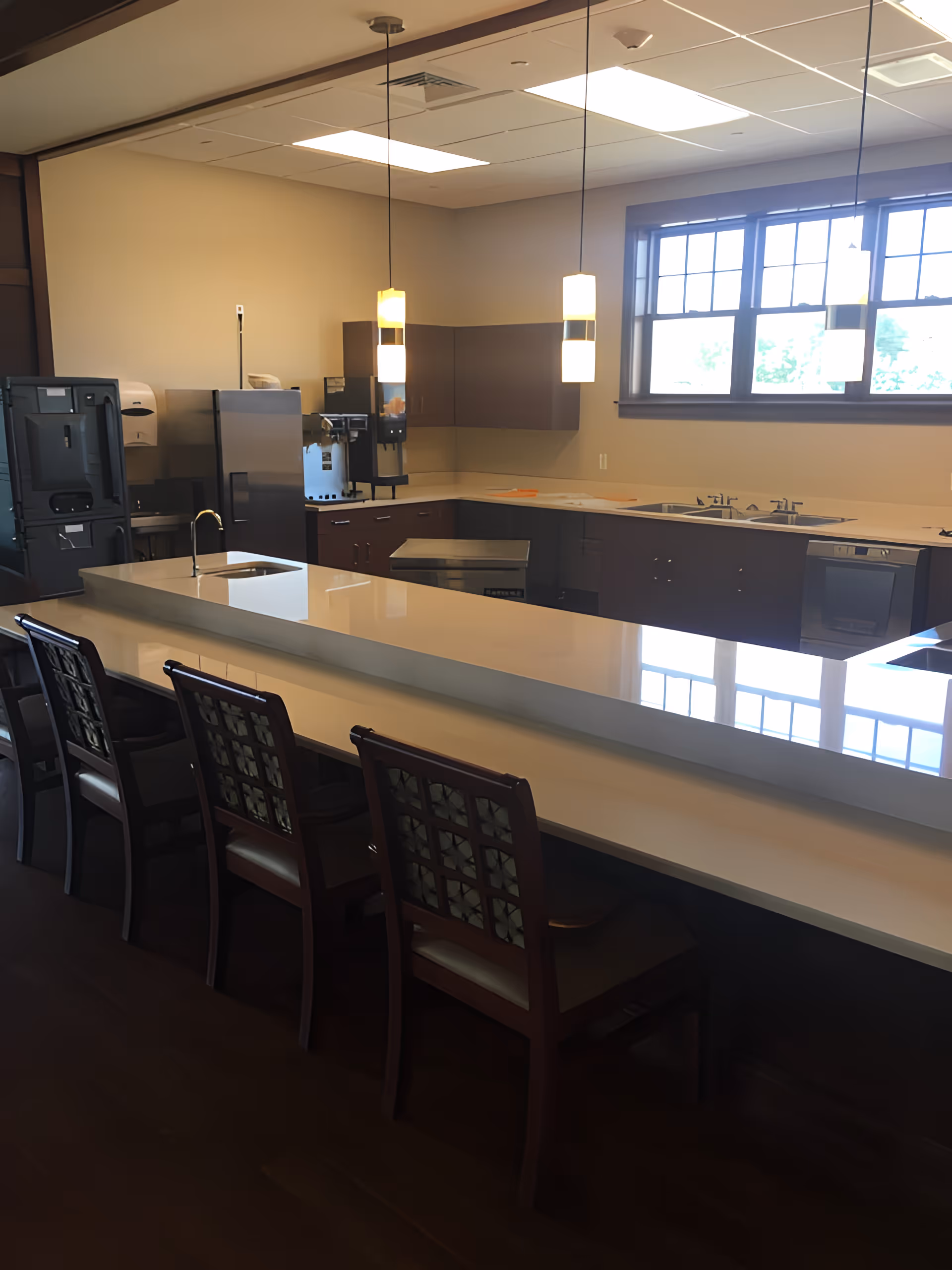 A modern kitchen area in Armstrong Memory Care Assisted Living featuring a long white countertop with an integrated sink and four wooden chairs with patterned backs. The kitchen has dark brown cabinets, a large window letting in natural light, pendant lights hanging from the ceiling, and various kitchen appliances including a refrigerator and coffee machine.