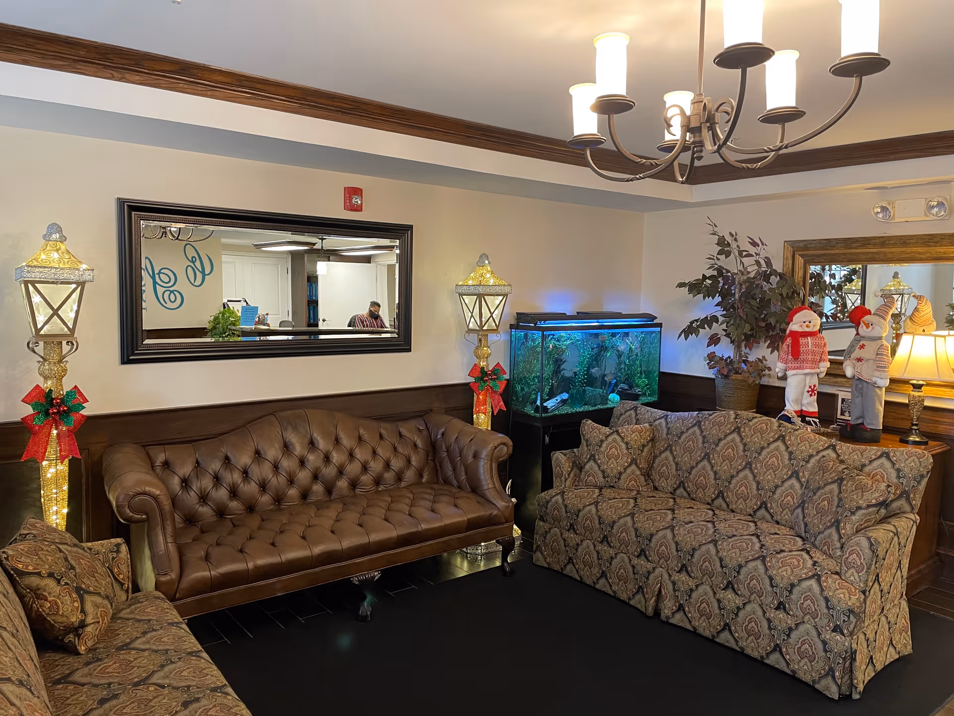 A cozy living room area with two patterned fabric sofas and one brown leather tufted sofa arranged around a black rug. There is a large fish tank on a stand in the corner, flanked by two decorative lamp posts wrapped with red bows and green accents. A large mirror hangs on the wall above the leather sofa, reflecting part of the room. A chandelier with multiple lights hangs from the ceiling. On a wooden side table near the fish tank, there are two snowman figurines dressed in winter clothing and a potted plant. The room has wood trim along the ceiling and walls.