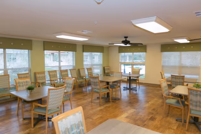 A bright dining room with multiple wooden tables and chairs arranged neatly on a wooden floor. Large windows with blinds allow natural light to fill the room. The walls are painted a light yellow-green color, and there is a ceiling fan and several ceiling lights.