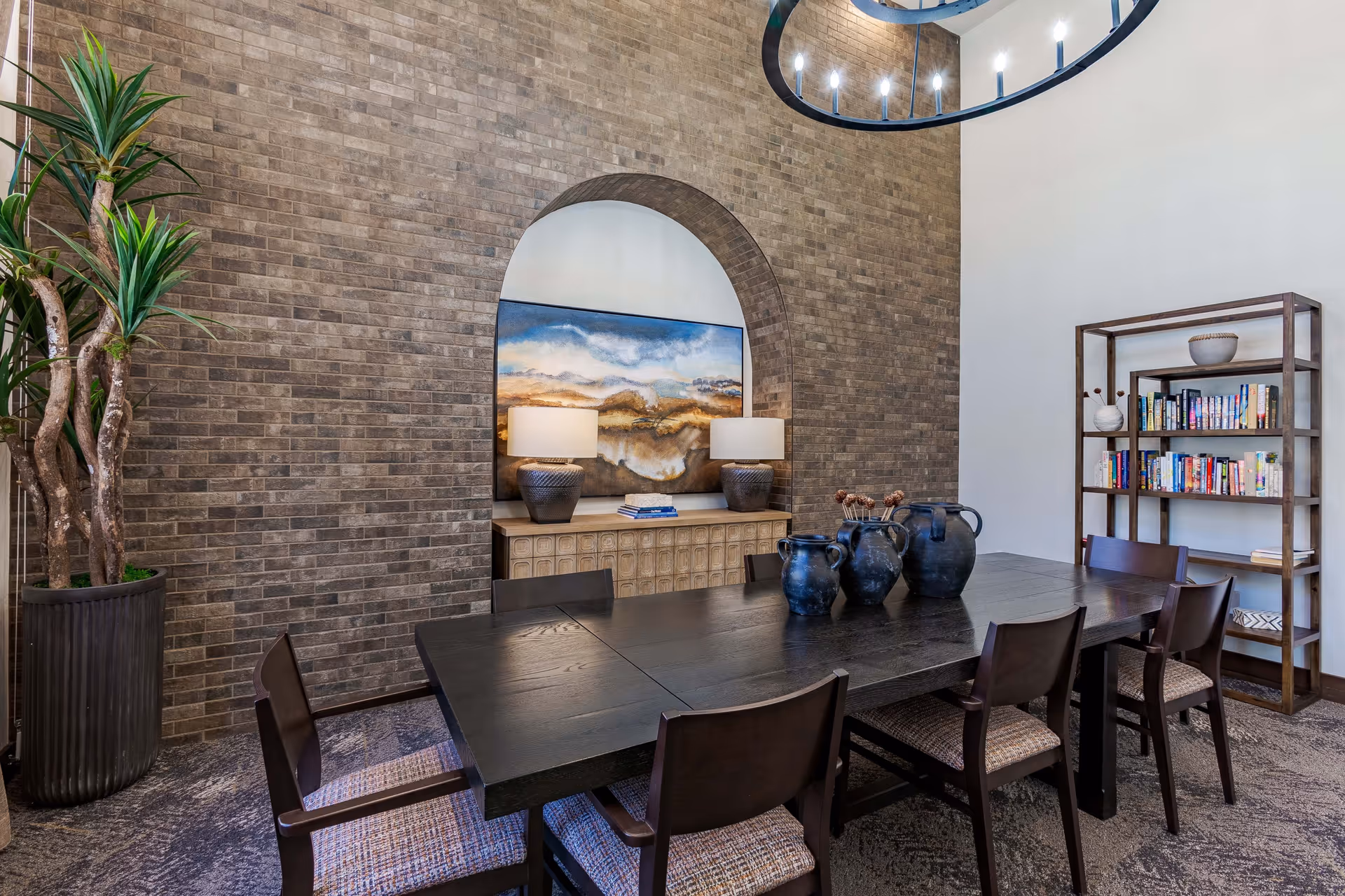 Communal dining room with a long dark wood table and chairs, decorative vases, an arched brick accent wall with artwork and lamps, a bookshelf and a tall potted plant.