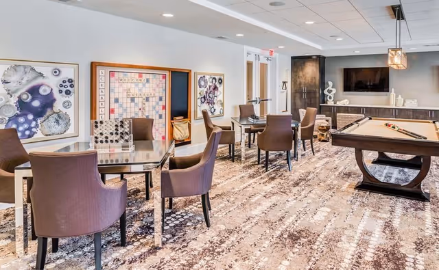 A bright and spacious game room with several tables and chairs, a large Scrabble board on the wall, abstract artwork, and a pool table with billiard balls and cues. The room has a patterned carpet and modern decor, including a TV mounted on the wall and decorative sculptures on a sideboard.