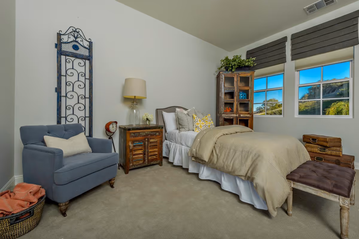 A cozy bedroom with a single bed covered in beige bedding and multiple pillows. Next to the bed is a rustic wooden nightstand with a lamp and a small flower vase. A blue upholstered armchair with a beige pillow is placed near the corner, next to a decorative metal wall piece. There is a wooden cabinet with glass doors and some decorative items inside, positioned between two windows with gray blinds. A small cushioned bench and a stack of vintage suitcases are at the foot of the bed. The room has beige carpet and light-colored walls.