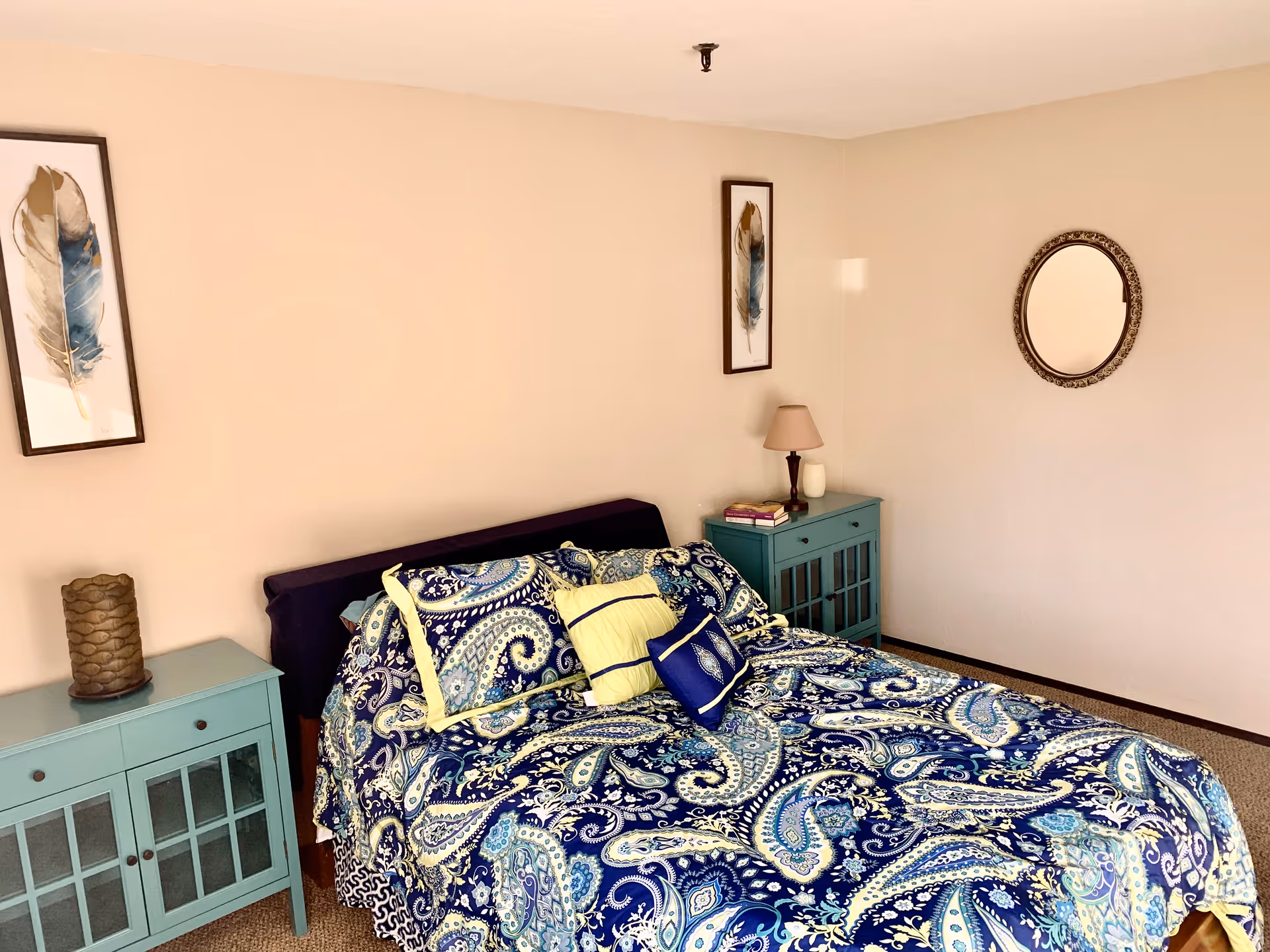 A bedroom with a bed covered in a blue and white paisley patterned bedspread and several pillows. There are two teal nightstands on either side of the bed, one with a lamp and books, and the other with a decorative item. The walls are beige with two framed feather artworks and a round mirror.
