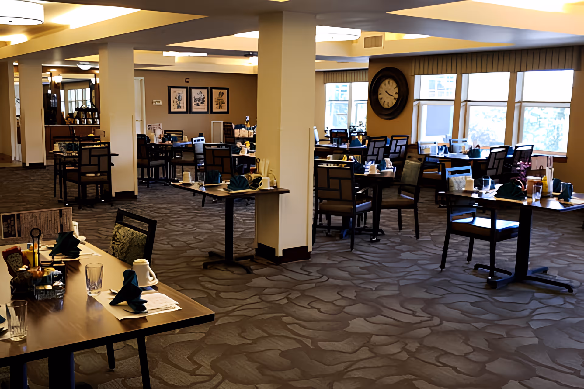 Spacious dining room with multiple set tables and chairs, a large wall clock, and windows letting in natural light.