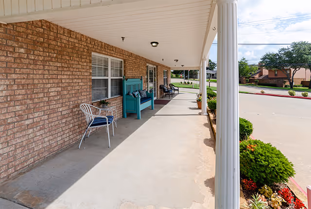 A covered outdoor walkway with a brick wall on one side and white columns supporting the roof. There are chairs and a teal bench with cushions along the wall. The walkway overlooks a paved driveway and landscaped bushes with flowers.