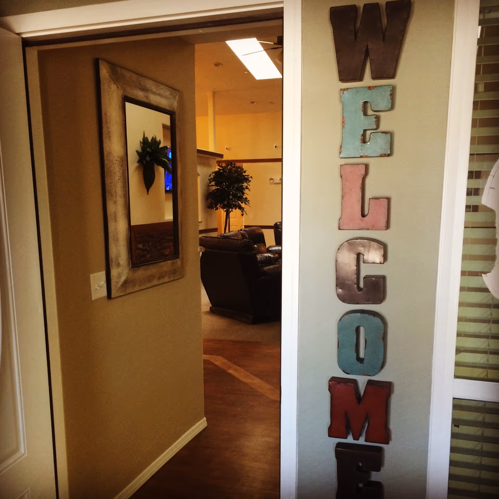 Interior view of a senior living facility showing a hallway with a large mirror on the left wall and a vertical decorative sign spelling 'WELCOME' on the right wall. In the background, there is a seating area with leather chairs and a potted plant under warm lighting.