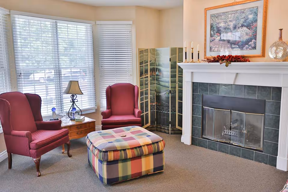 A cozy living room area with two red upholstered armchairs, a plaid ottoman, a wooden side table with a lamp and decorative items, a fireplace with green tiles and white mantel, and a framed painting above the fireplace. There is also a decorative folding screen in the corner and large windows with white blinds letting in natural light.