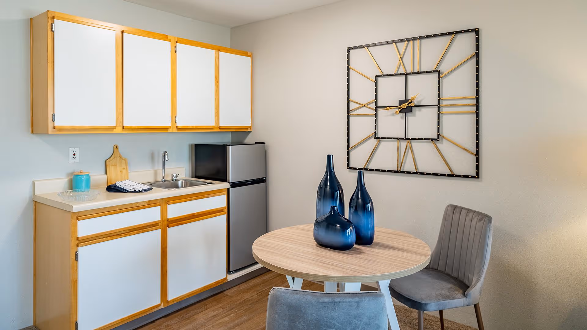A small kitchen area with light wood and white cabinets, a stainless steel sink, a mini refrigerator, and a round wooden table with three blue decorative vases. Two gray upholstered chairs are positioned around the table, and a large decorative wall clock with a black frame and gold Roman numerals hangs on the wall.