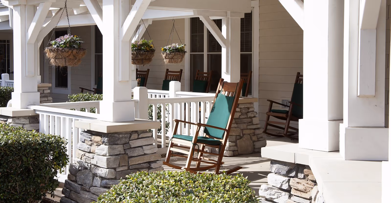 Front porch with wooden rocking chairs, hanging flower baskets, white columns and stone pillars overlooking trimmed shrubs.