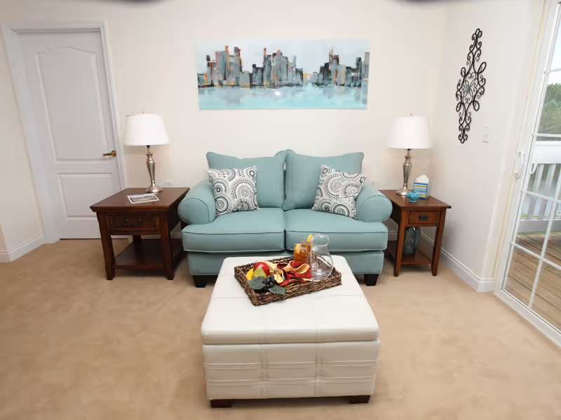 A cozy living room with a light blue loveseat adorned with two patterned pillows. On either side of the loveseat are wooden side tables with matching lamps. A white ottoman sits in front of the loveseat with a decorative tray holding a glass pitcher, a glass of iced tea, and some fruit. A cityscape painting hangs on the wall above the loveseat, and a sliding glass door is visible on the right side, leading to an outdoor balcony.
