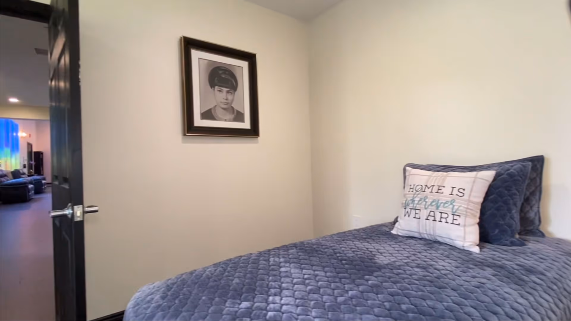 Small bedroom with a blue quilted bed and decorative pillow, a framed portrait on the wall, and an open door showing the living area.