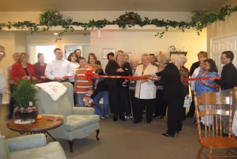 A group of people gathered indoors for a ribbon-cutting ceremony. Two women in the center are holding large scissors cutting a red ribbon. The room has light-colored walls, green plants decorating the top edges, and comfortable seating including armchairs and wooden chairs. The atmosphere appears celebratory and communal.