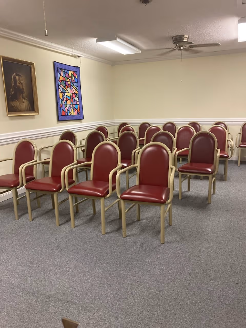 A room with multiple rows of maroon cushioned chairs with beige frames arranged facing forward. The walls are light-colored with white trim, and there is a framed portrait of Jesus and a colorful abstract wall hanging on one wall. The ceiling has fluorescent lights and a ceiling fan.