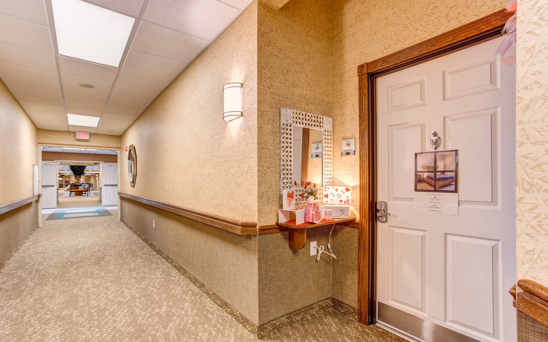 A well-lit hallway in a retirement facility with beige patterned walls and carpeted floor. On the right side, there is a white door with a wooden frame, decorated with a small picture and a note. Next to the door is a small wall-mounted shelf with a mirror above it, holding a lamp and some decorative items. At the end of the hallway, double doors open into a larger common area with seating and plants.