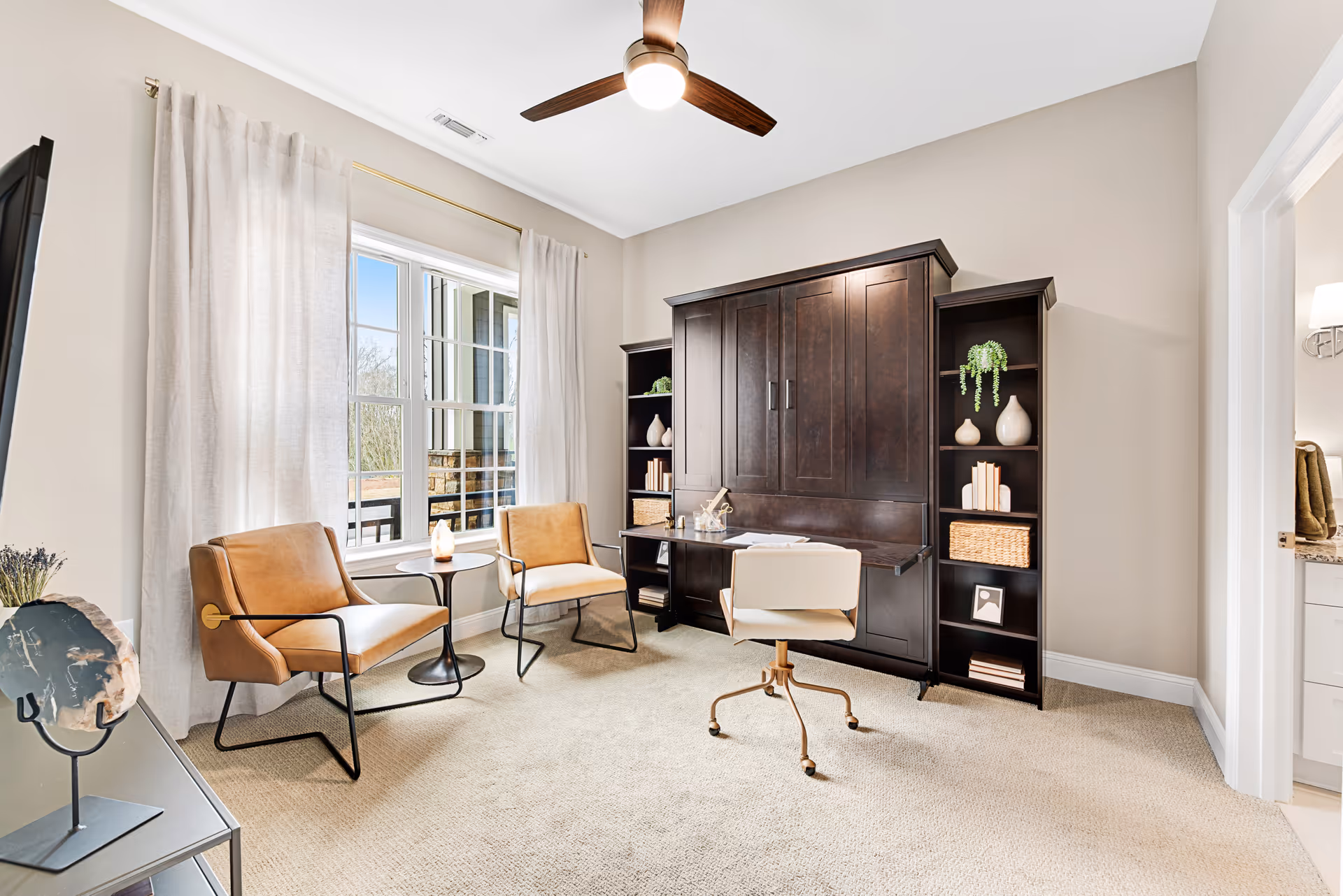 A bright room with beige walls and carpet featuring two tan leather chairs with black metal frames, a small round black side table between them, a large window with white curtains, a ceiling fan with a light, and a dark wood Murphy bed with built-in shelves on either side. A white swivel chair is positioned in front of the Murphy bed, which is folded up to reveal a desk surface. The room opens to a bathroom visible on the right side.