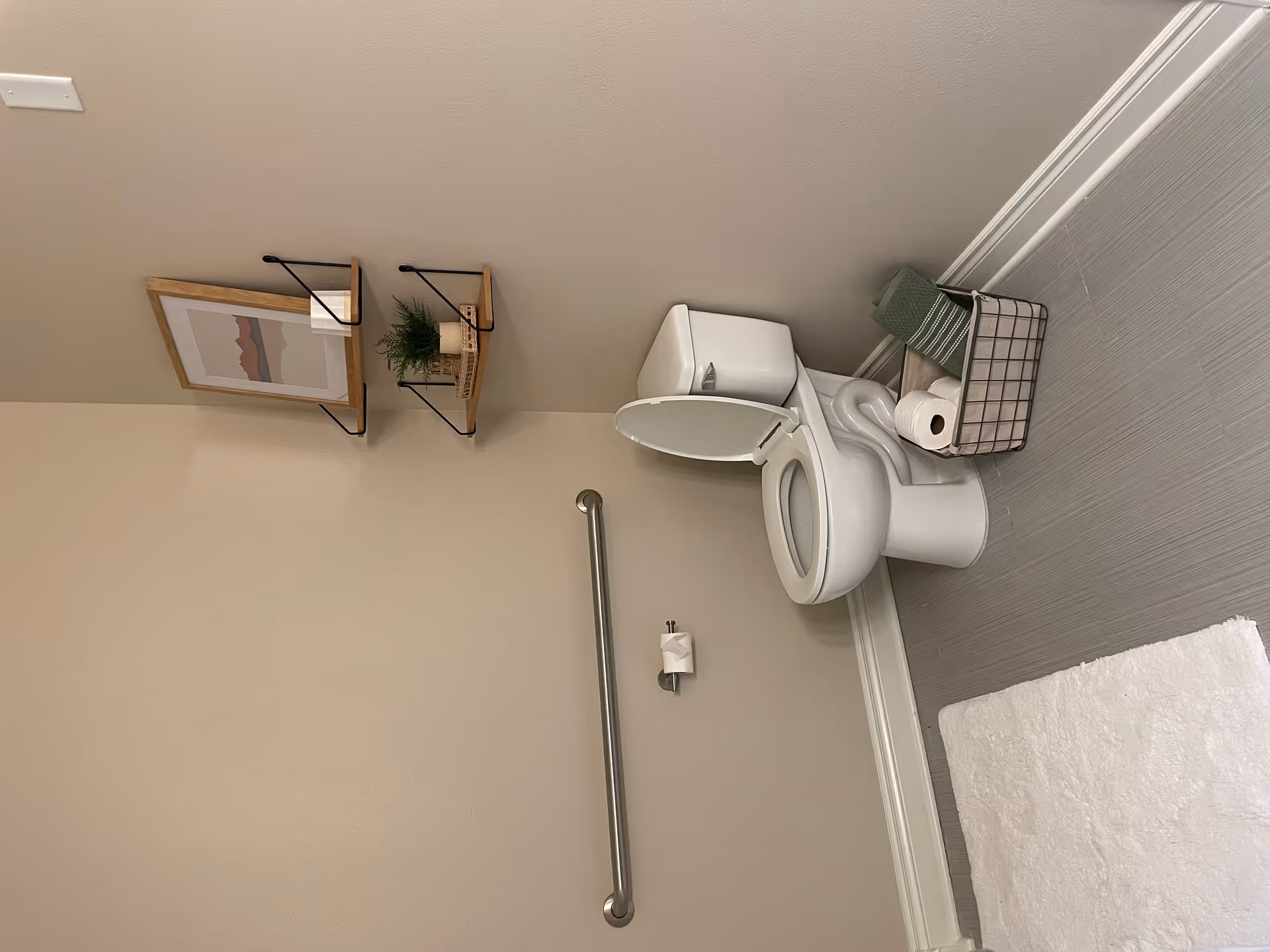Small accessible bathroom with a toilet (seat up), a wall grab bar, two decorative wall shelves, and a basket holding towels and toilet paper.