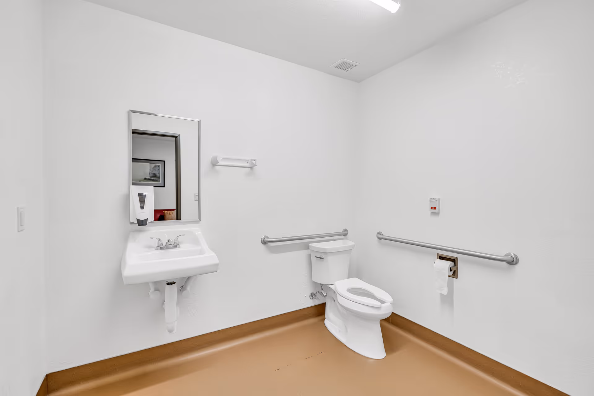 Accessible institutional bathroom with a wall-mounted sink and mirror, toilet, and grab bars on white walls and a brown floor.
