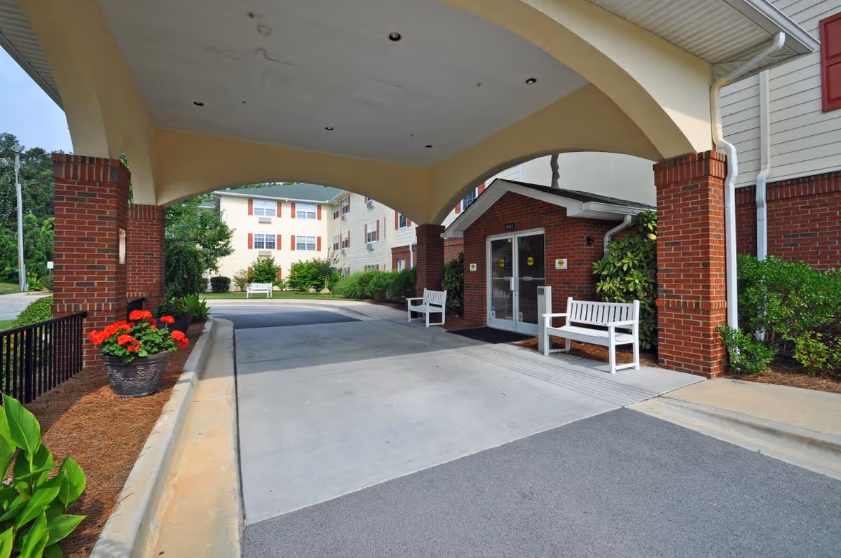 Covered driveway and entrance of a brick retirement community building with white benches, planters, and a porte-cochere.