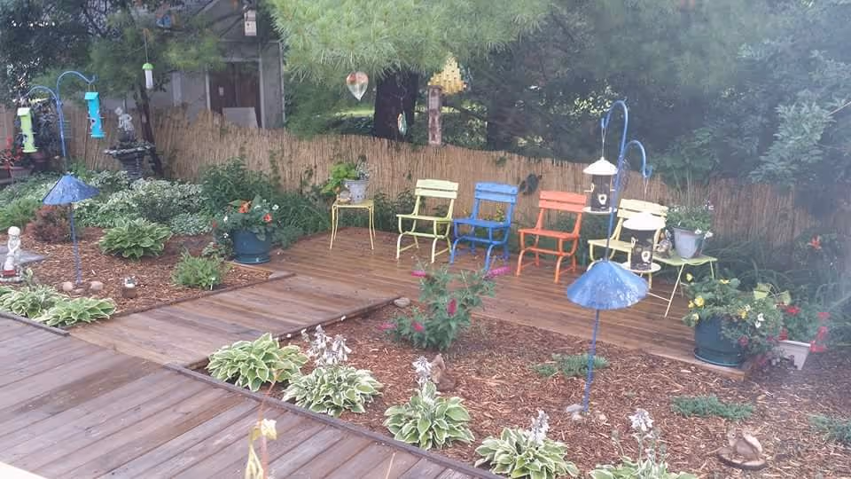 Outdoor garden area with a wooden deck featuring colorful metal chairs in yellow, blue, and orange. The garden has various green plants, flowers, bird feeders, and decorative elements surrounded by a wooden fence and trees in the background.